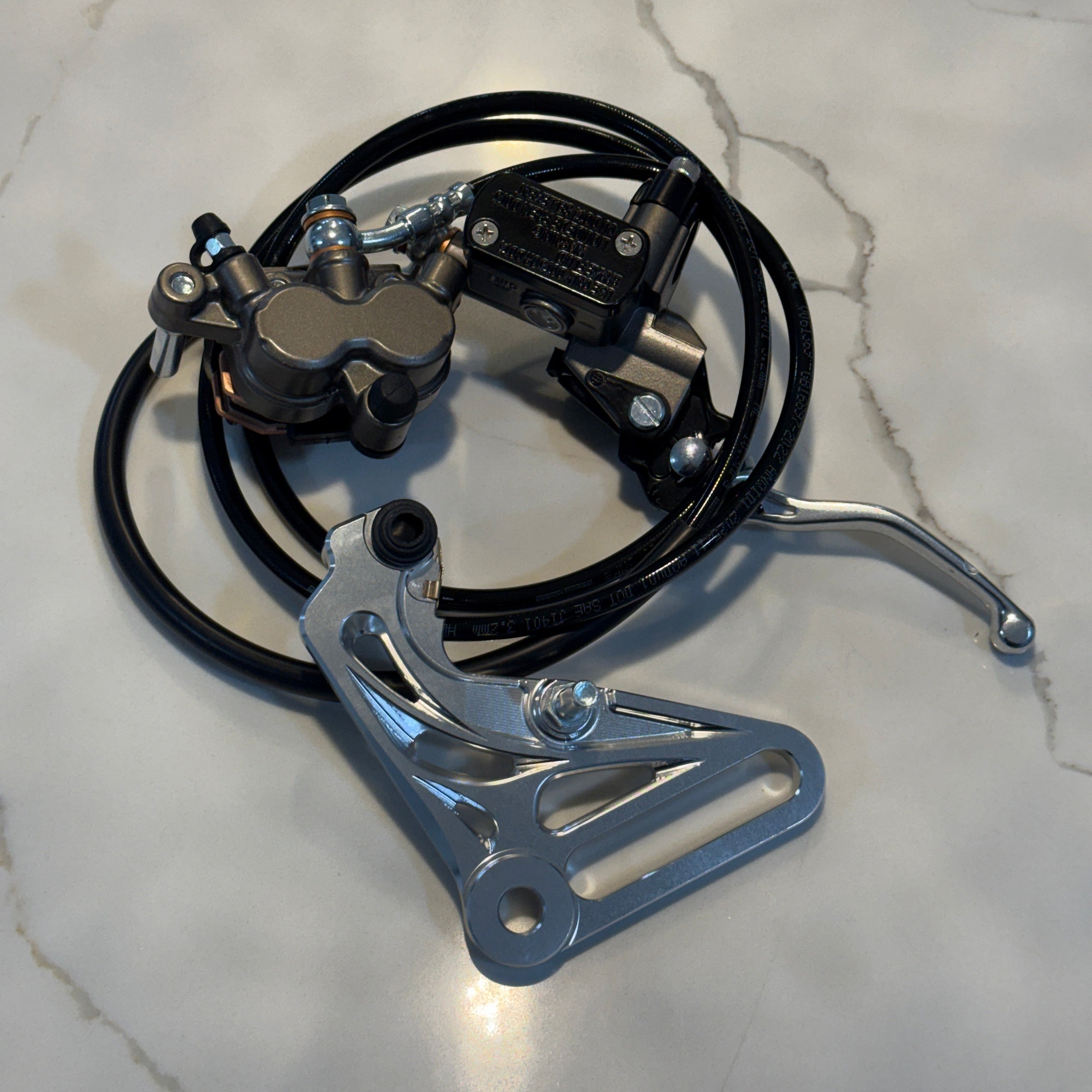 A motorcycle brake and clutch assembly with black hoses and silver metal components, photographed on a light marble surface.